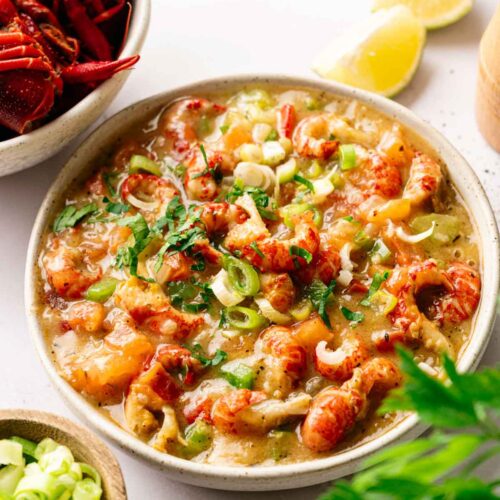 A bowl of Crawfish Etouffee garnished with chopped green onions and parsley, with lemon wedges and a side of crawfish and sliced green onions nearby.