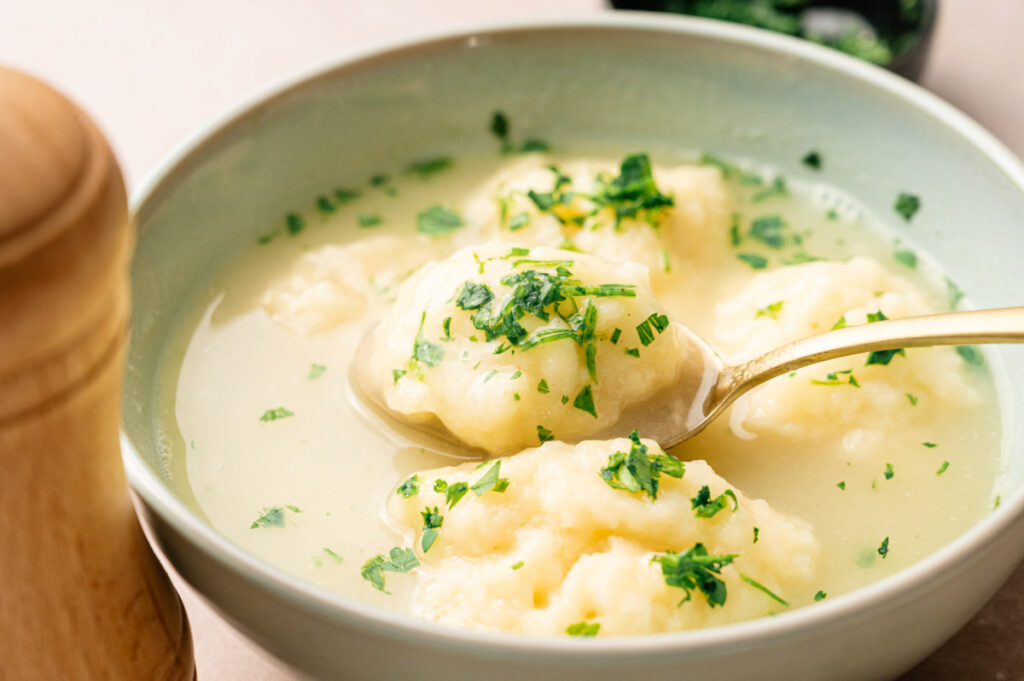 A bowl of clear soup with fluffy dumplings, garnished with chopped parsley, with a spoon lifting one delicious dumpling.