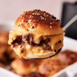 A close-up of an Air Fryer Hamburger Slider with minced beef and melted cheese in a sesame seed bun, held above a tray of more sliders.