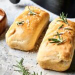 Two rectangular loaves of Air Fryer Sourdough Focaccia, topped with coarse salt and rosemary sprigs, are placed on a light surface beside a small wooden bowl of salt.