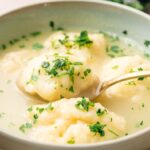 A bowl of soup with three dumplings, topped with chopped fresh herbs, with a spoon lifting one dumpling from the savory broth.