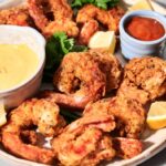 A plate of crispy fried shrimp with lemon wedges, parsley, and two dipping sauces—one yellow and one red—served in small bowls.