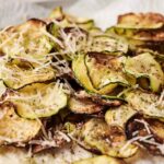 Sliced and baked zucchini chips reminiscent of classic Fried Zucchini, topped with shredded cheese and pepper, served on a white surface.