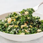 A white bowl filled with Kale Salad with Lemon and Parmesan, topped with sliced almonds and grated cheese, with a fork lifting a portion.