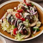 Three grilled Lamb Kebabs on flatbread with shredded lettuce, cherry tomatoes, red onion, lime wedges, and drizzled creamy sauce on a brown plate.