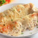 Two creamy white chicken enchiladas topped with melted cheese and sprinkled with chopped herbs are served on a white plate.