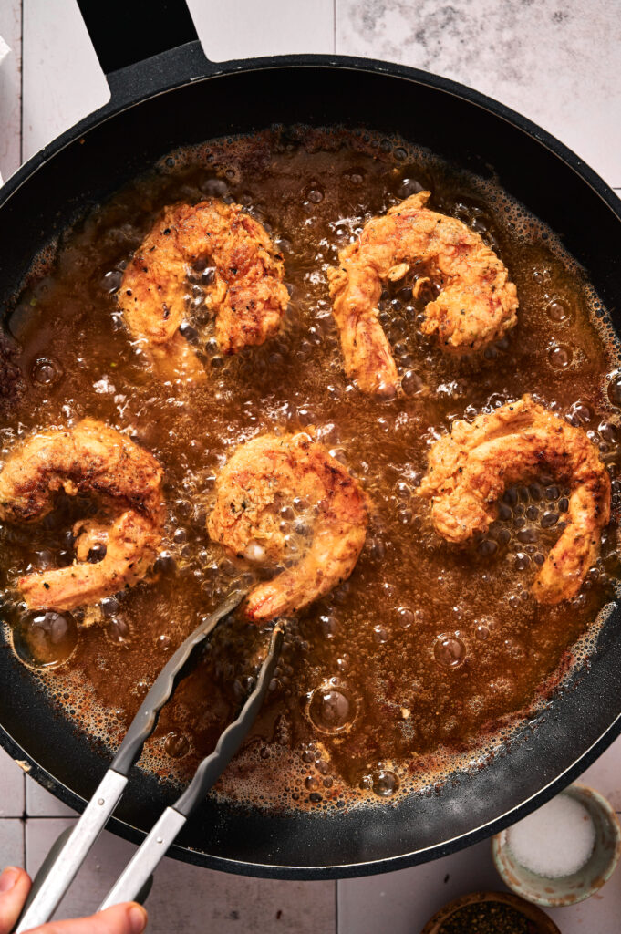 Four fried shrimp are being crisped to perfection in hot oil in a black pan. A hand holding metal tongs is turning one of the golden shrimp for even frying.