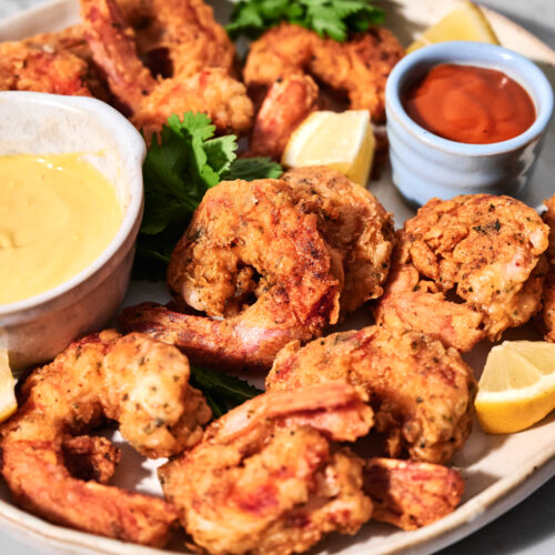A plate of crispy fried shrimp served with lemon wedges, dipping sauces, and a parsley garnish on a light-colored surface.