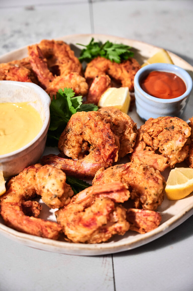 A plate of crispy fried shrimp served with lemon wedges, dipping sauces, and a parsley garnish on a light-colored surface.