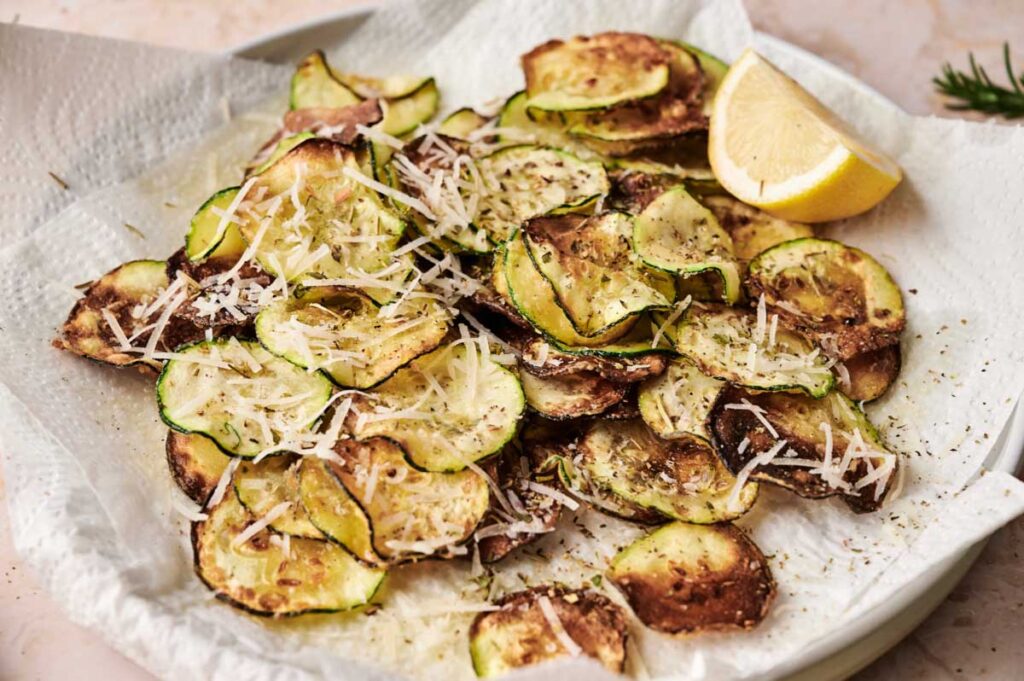 A plate of baked zucchini chips, inspired by classic Fried Zucchini, topped with grated cheese and served on a paper towel with a lemon wedge on the side.