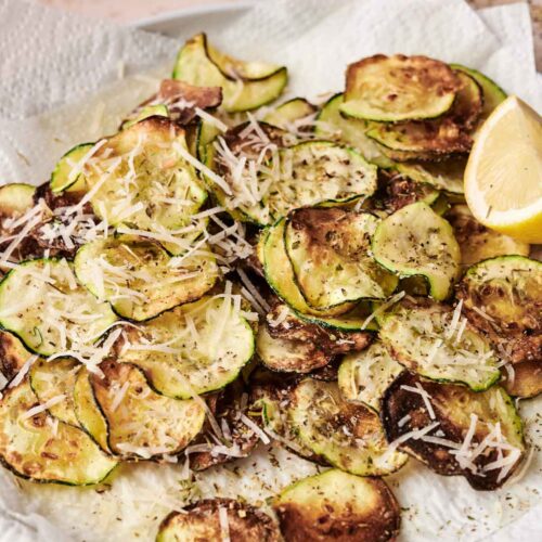 A plate of baked zucchini chips reminiscent of classic fried zucchini, topped with shredded parmesan cheese and herbs, served on a paper towel with a lemon wedge.