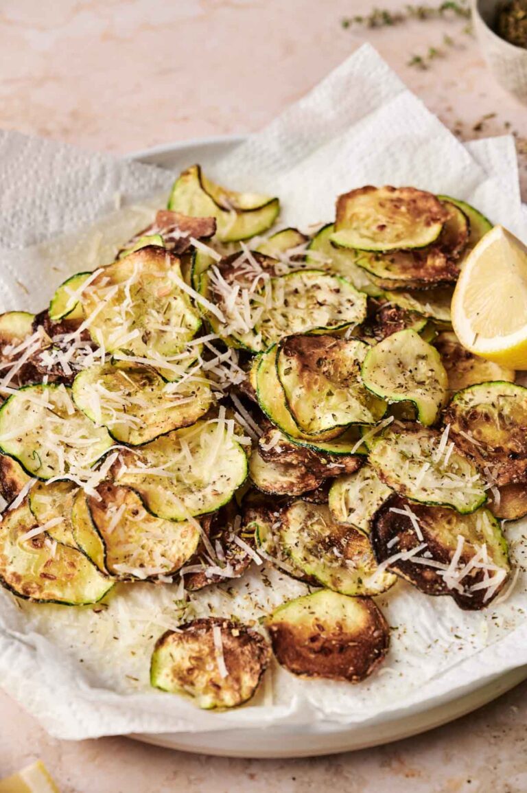 A plate of baked zucchini chips reminiscent of classic fried zucchini, topped with shredded parmesan cheese and herbs, served on a paper towel with a lemon wedge.