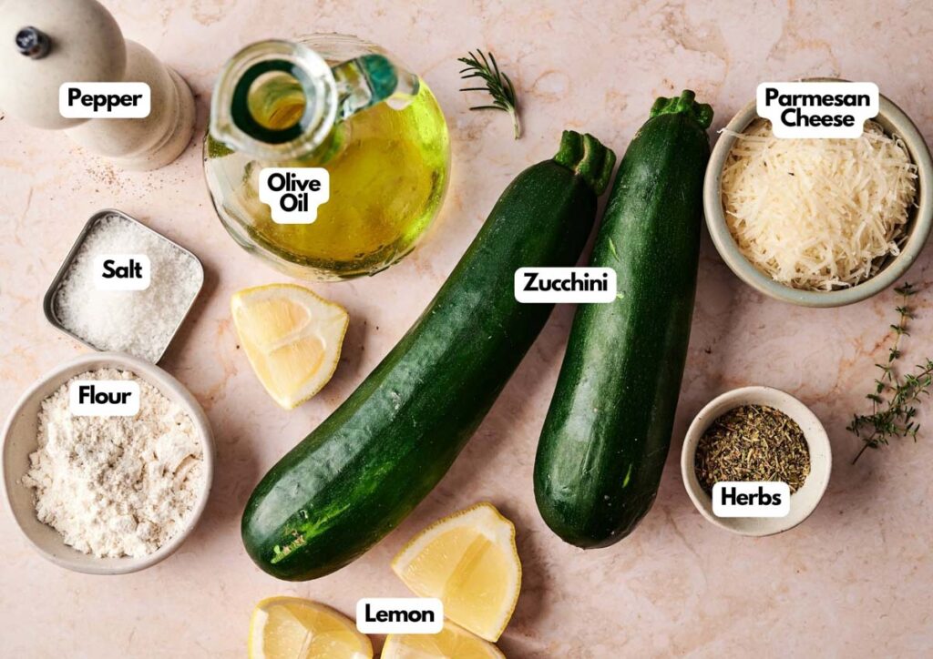 Ingredients for Fried Zucchini, including two zucchinis, olive oil, parmesan cheese, flour, salt, pepper, herbs, and lemon wedges arranged on a countertop.