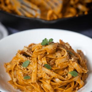 A white bowl of Hidden Veggie Pasta in a creamy orange sauce, garnished with fresh herbs and breadcrumbs, with a skillet of more pasta in the background.