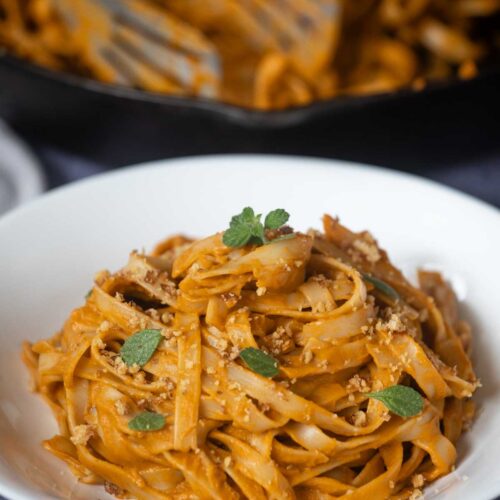 A white bowl of Hidden Veggie Pasta in a creamy orange sauce, garnished with fresh herbs and breadcrumbs, with a skillet of more pasta in the background.