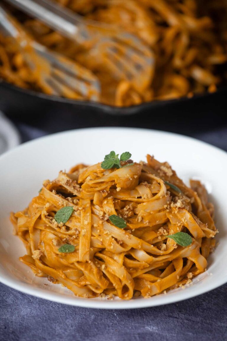 A white bowl of Hidden Veggie Pasta in a creamy orange sauce, garnished with fresh herbs and breadcrumbs, with a skillet of more pasta in the background.