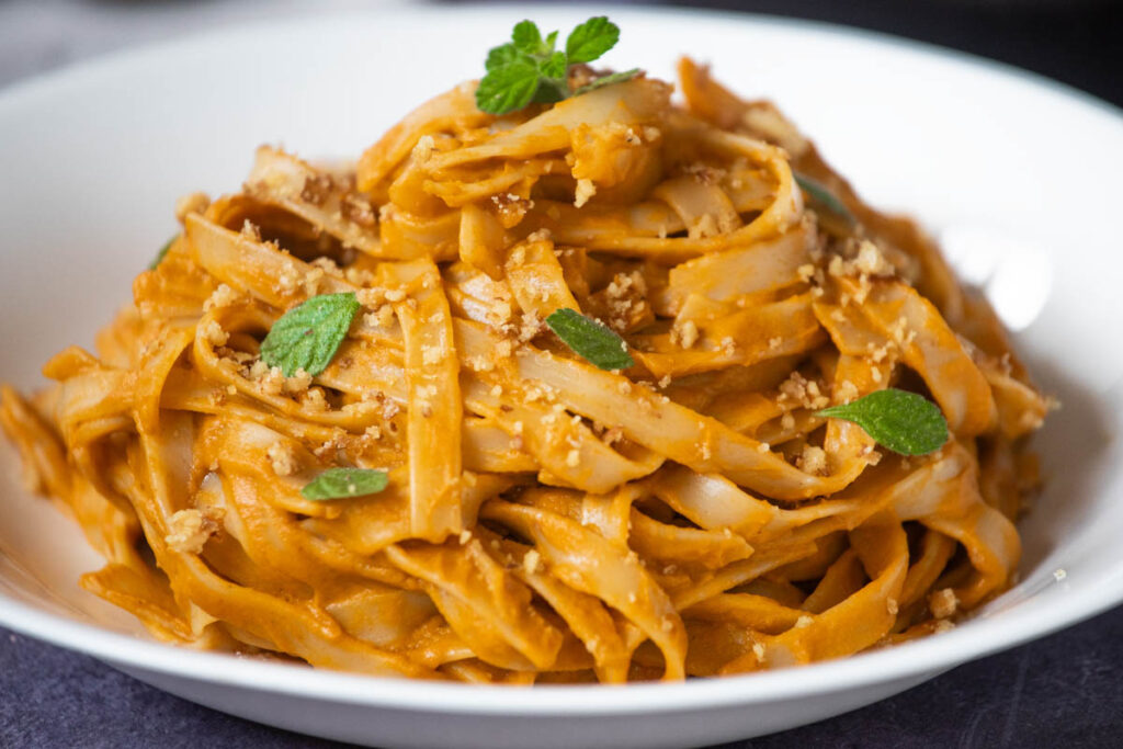 A white bowl filled with Hidden Veggie Pasta—wide noodles coated in a creamy orange sauce, garnished with chopped nuts and fresh herbs.