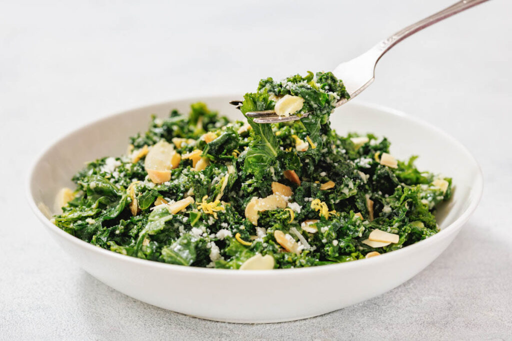 A white bowl filled with Kale Salad with Lemon and Parmesan, topped with sliced almonds, grated cheese, and lemon zest, with a fork lifting a portion.
