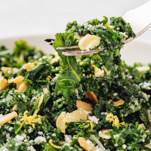 A close-up of a fork lifting Kale Salad with Lemon and Parmesan, sliced almonds, grated cheese, and lemon zest from a white bowl.