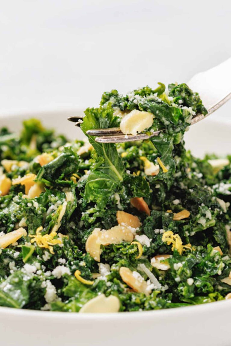 A close-up of a fork lifting Kale Salad with Lemon and Parmesan, sliced almonds, grated cheese, and lemon zest from a white bowl.