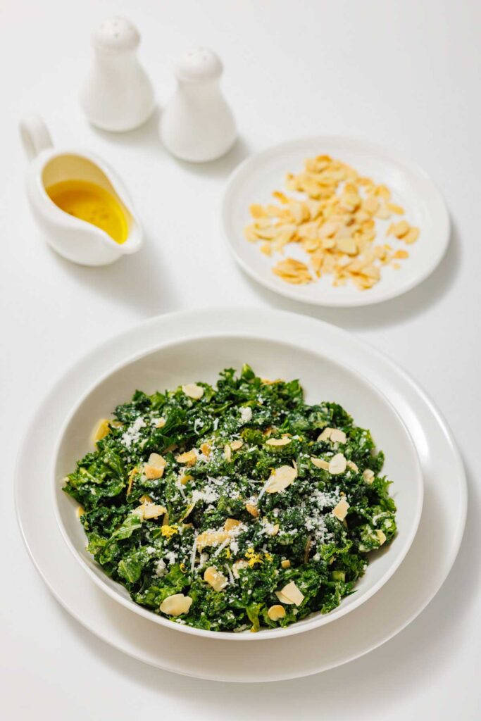 A bowl of Kale Salad with Lemon and Parmesan is topped with sliced almonds and grated cheese, with a plate of almonds, a small jug of dressing, and salt and pepper shakers in the background.