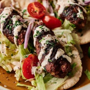 Grilled lamb kebabs on flatbread with shredded lettuce, cherry tomatoes, sliced red onions, and a drizzle of white sauce.
