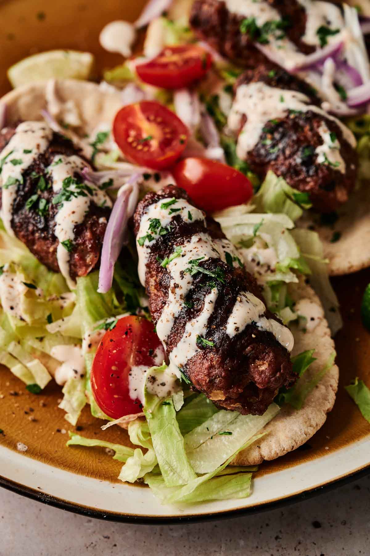 Grilled lamb kebabs on flatbread with shredded lettuce, cherry tomatoes, sliced red onions, and a drizzle of white sauce.