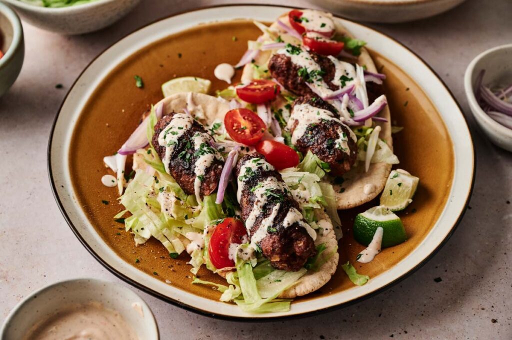A plate with pita bread topped with succulent lamb kebabs, shredded lettuce, cherry tomatoes, red onions, and a drizzle of white sauce, garnished with herbs and lime wedges.