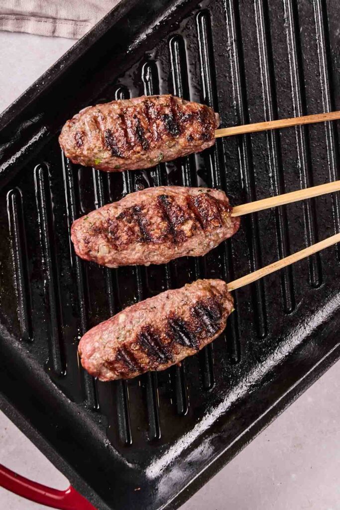 Three juicy lamb kebabs on wooden skewers are cooking on a ridged grill pan.