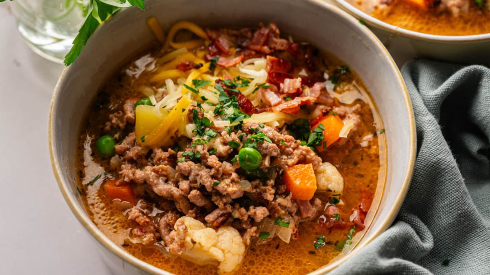 A bowl of soup with ground meat, peas, carrots, cauliflower, shredded cheese, chopped herbs, and bacon pieces, served with a napkin on the side.