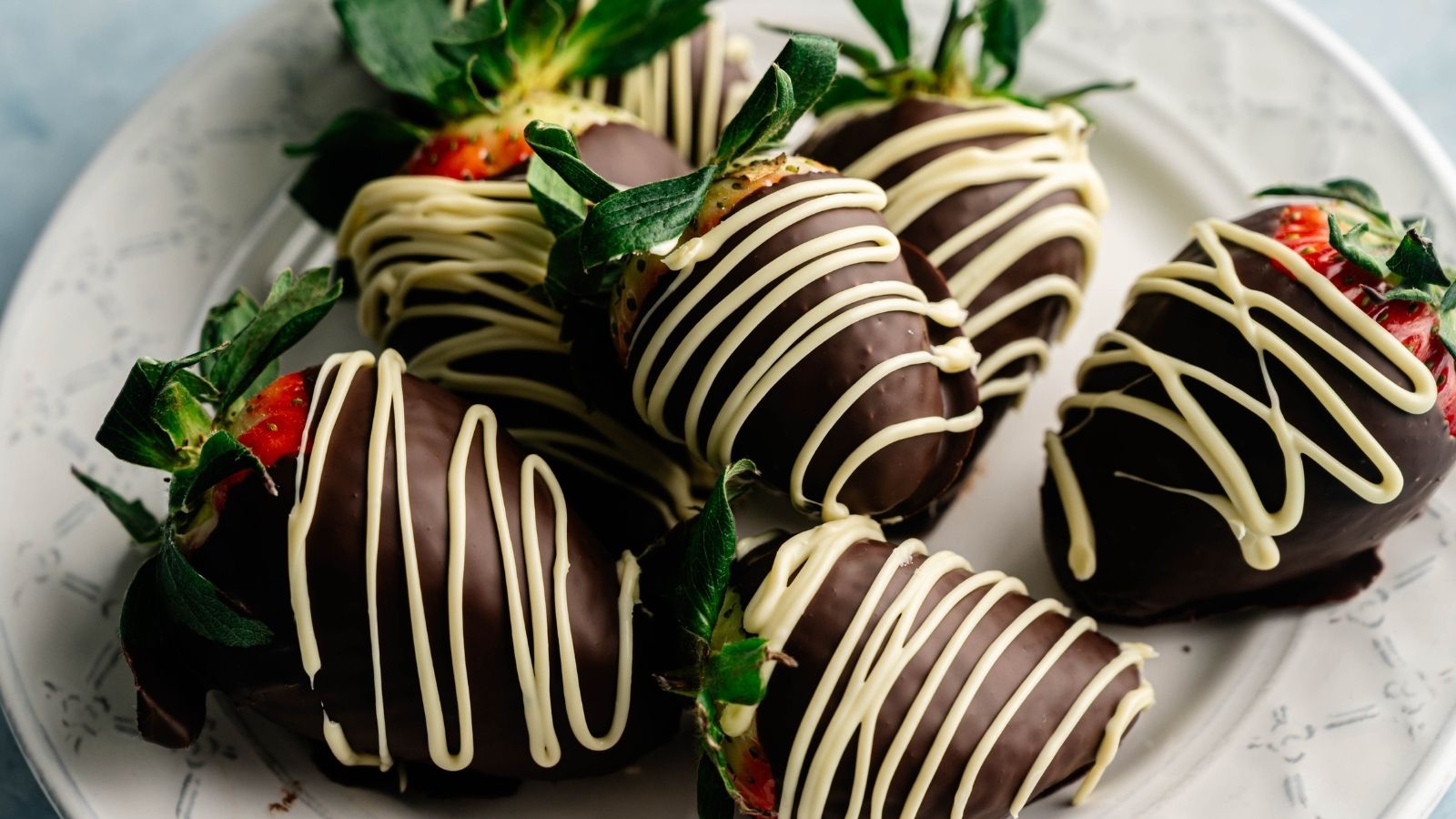 Six strawberries dipped in dark chocolate and drizzled with white chocolate are arranged on a white plate.