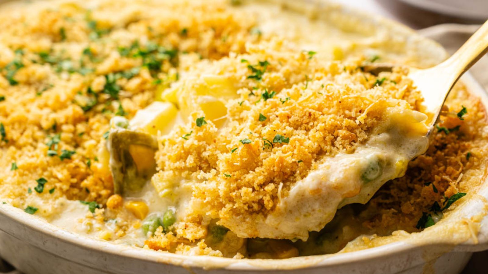 A spoon scoops a portion of creamy vegetable casserole topped with golden breadcrumbs and chopped herbs.