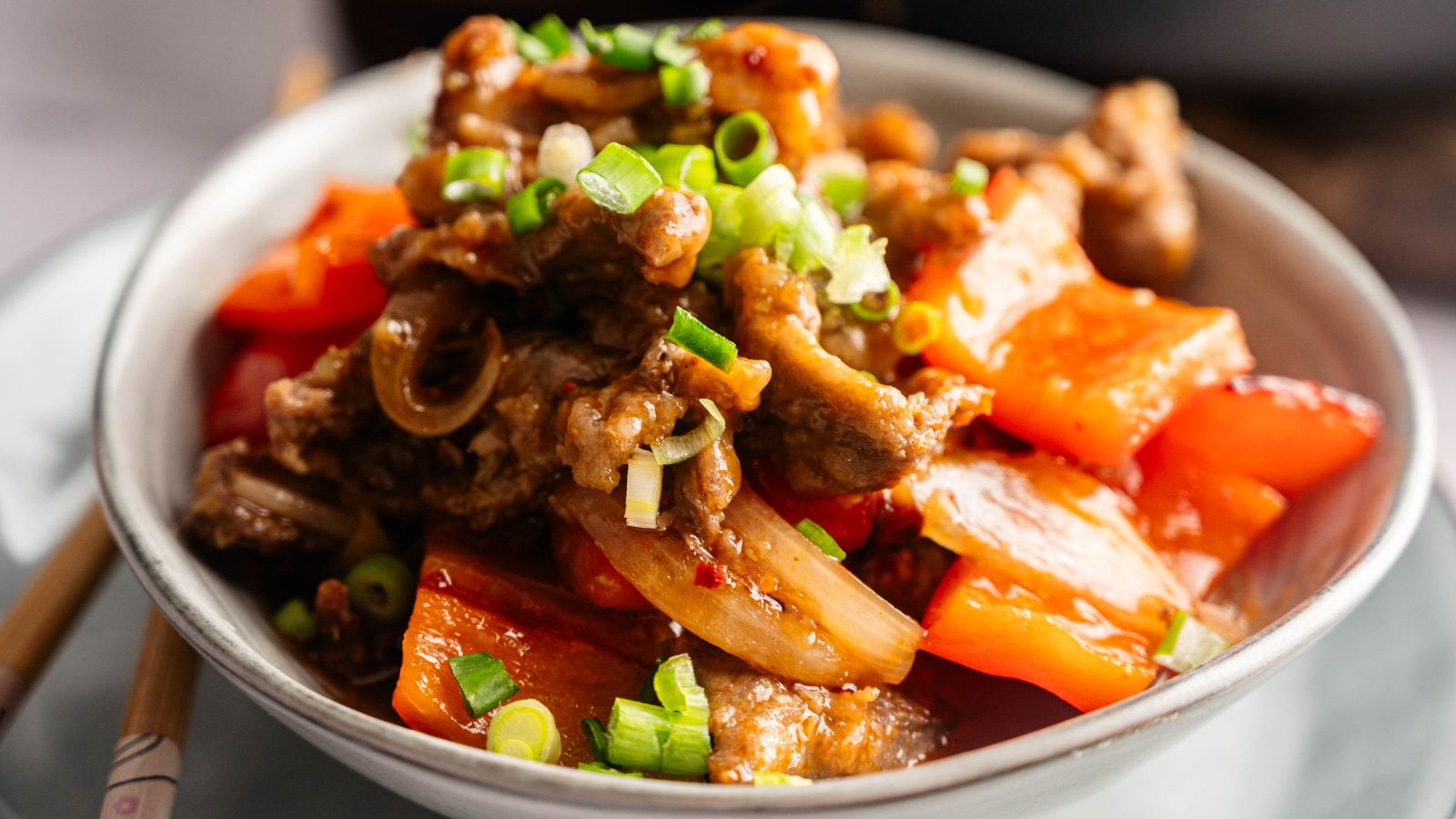 A bowl of stir-fried beef with red bell peppers, onions, and chopped green onions, served on a white plate.