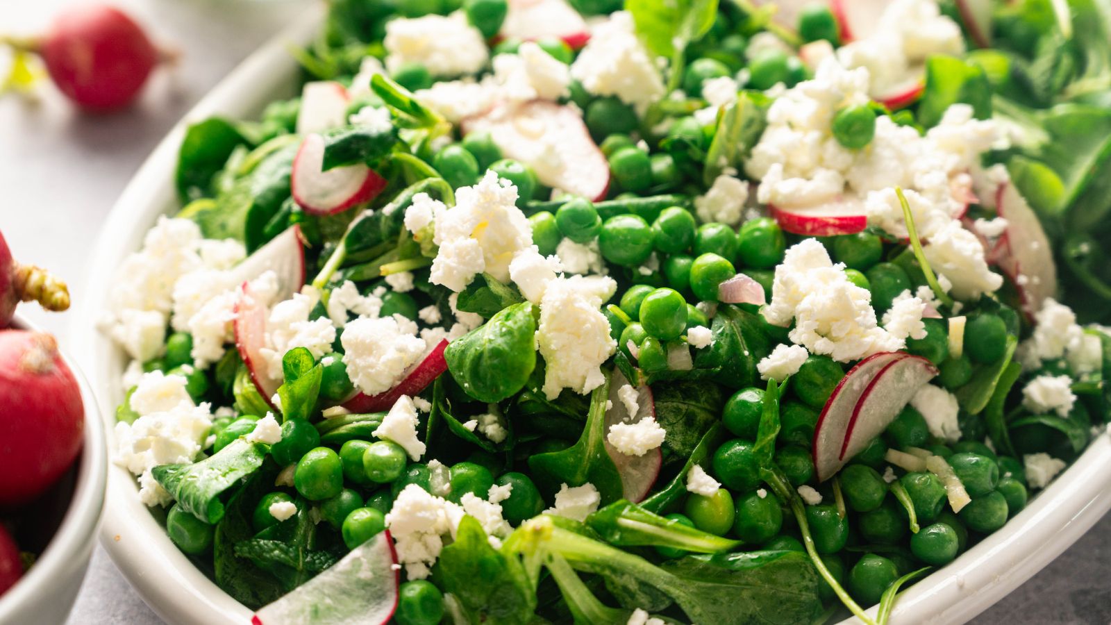 A close-up of a salad with green peas, leafy greens, sliced radishes, and crumbled feta cheese on a white plate.