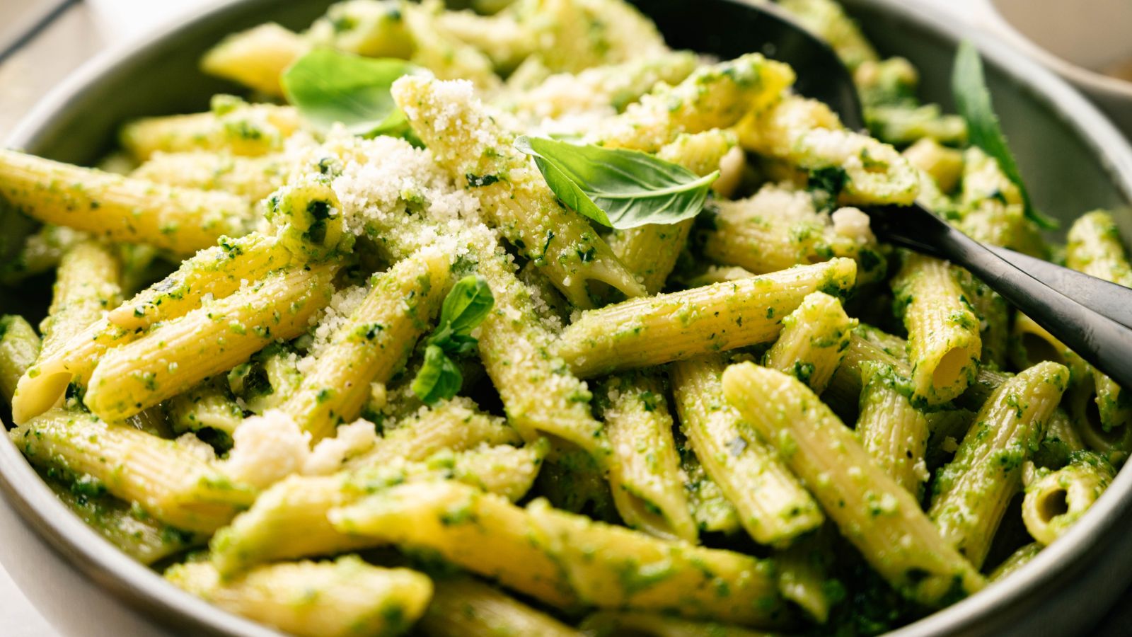 A bowl of penne pasta with green pesto sauce, topped with grated cheese and fresh basil leaves, with a black serving spoon.