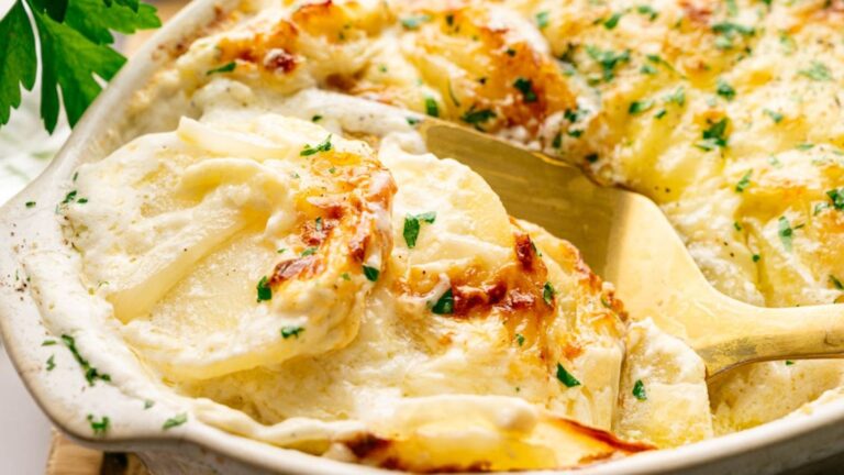 A close-up of a baked dish of creamy scalloped potatoes, topped with melted cheese and garnished with chopped parsley, with a serving spoon lifting a portion.