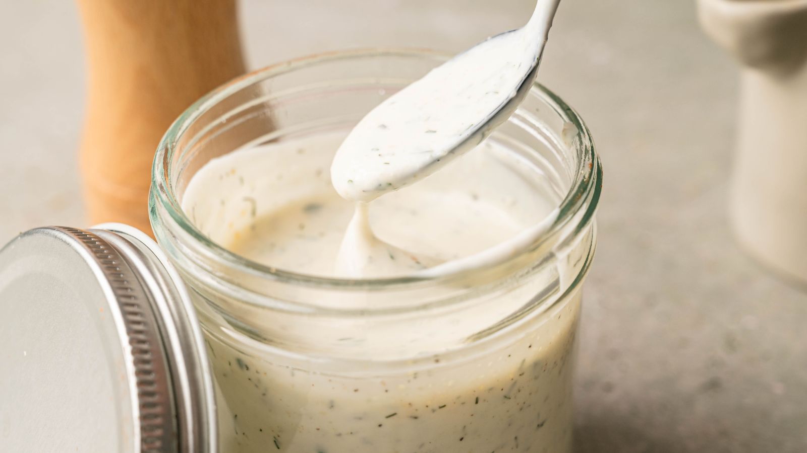 A spoon being dipped into a glass jar filled with creamy white dressing, with herbs visible throughout the mixture.