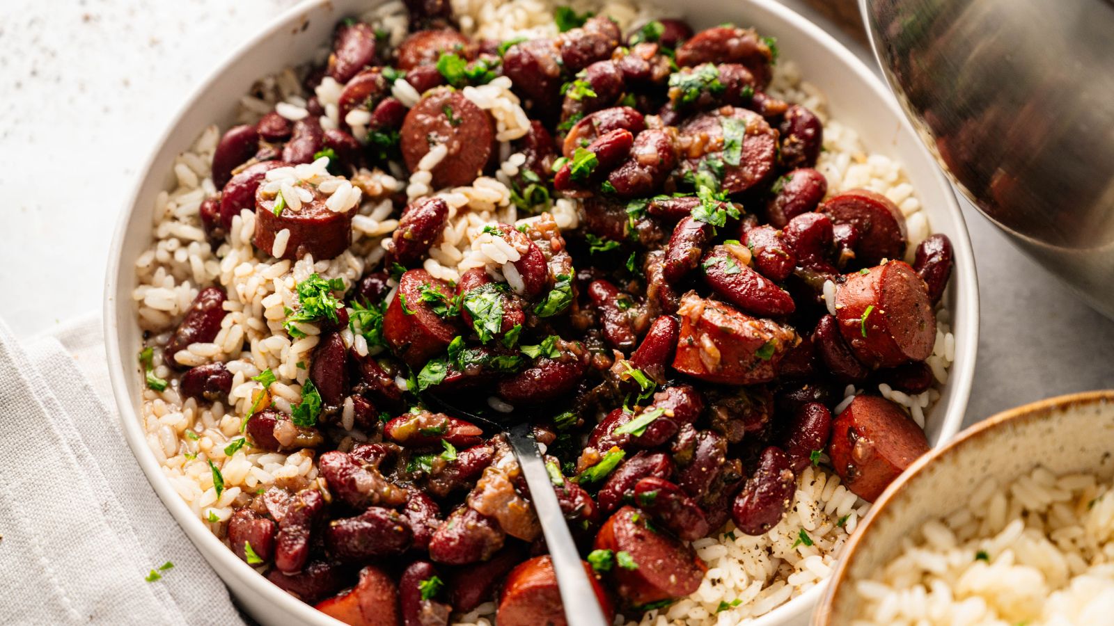 A bowl of white rice topped with kidney beans, sliced sausage, and chopped herbs, with a fork resting inside.