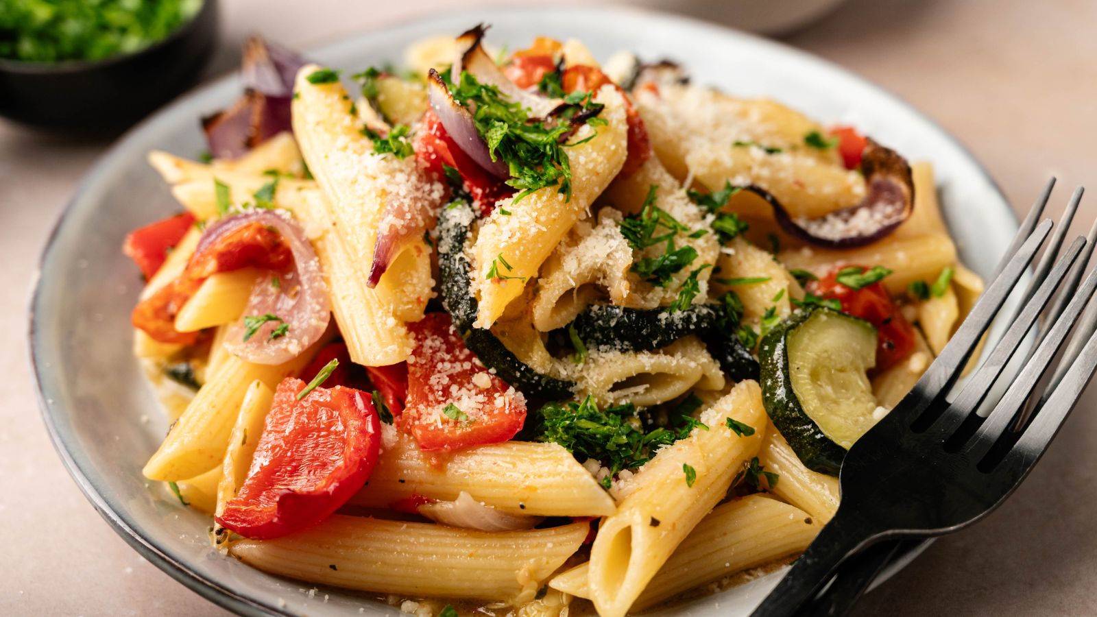 A plate of penne pasta with roasted vegetables, sprinkled with grated cheese and chopped herbs, with a black fork on the side.