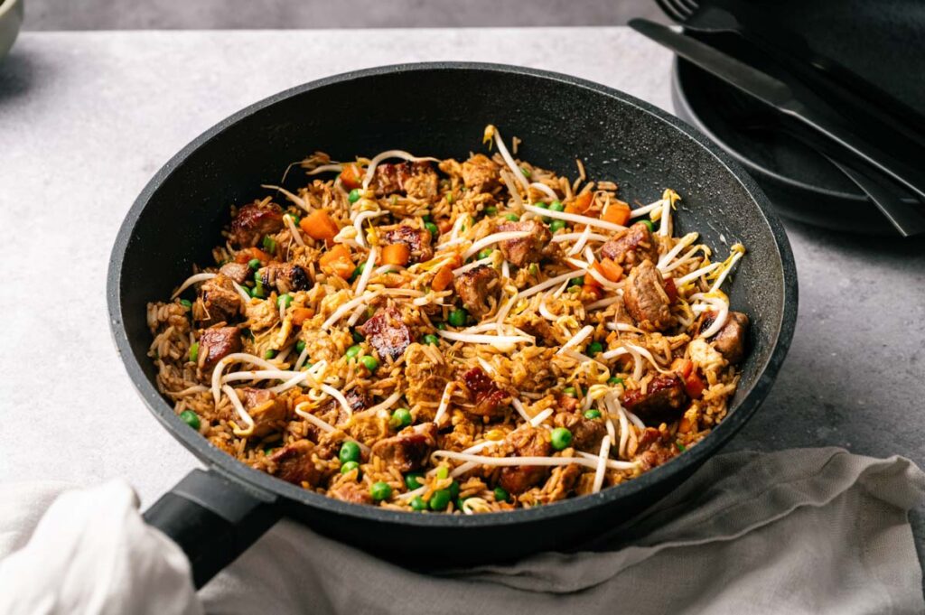 A skillet filled with Pork Fried Rice, featuring diced meat, carrots, peas, and bean sprouts on a gray surface with black cutlery and plates in the background.