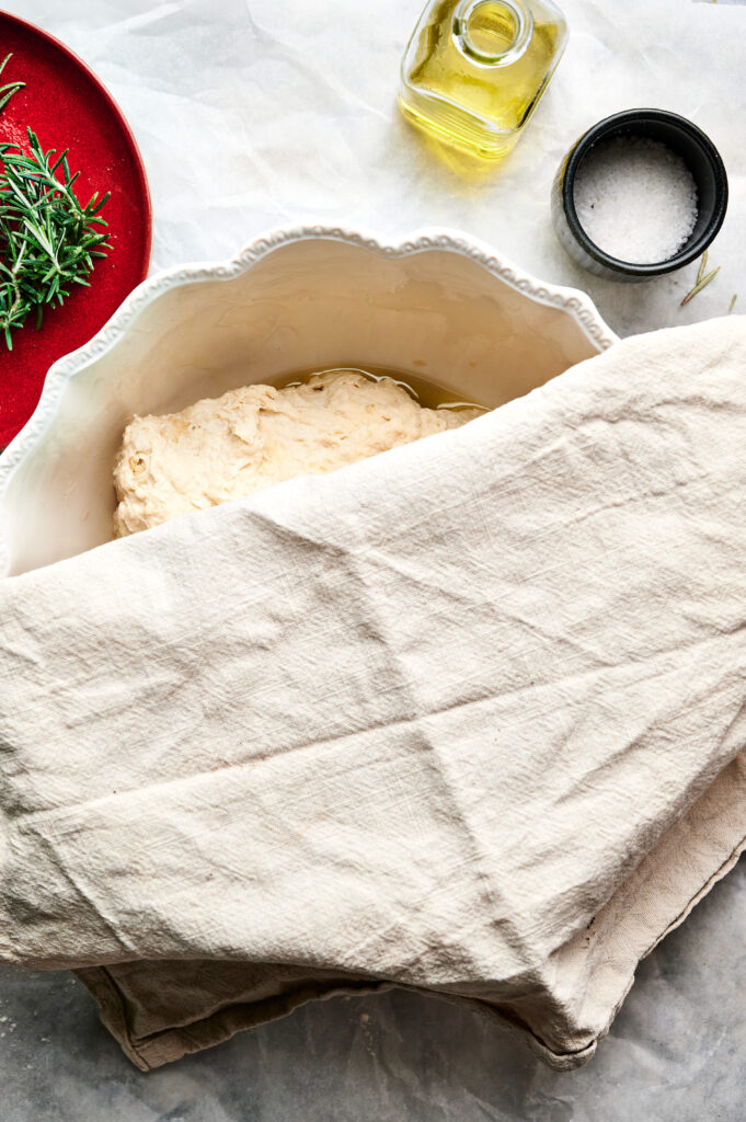 A bowl of dough for Air Fryer Sourdough Focaccia rests covered on the countertop, surrounded by a sprig of rosemary, a red plate, a bottle of olive oil, and a small bowl of salt.