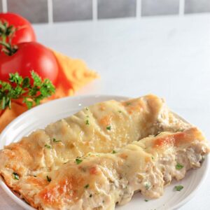 Two baked White Chicken Enchiladas topped with melted cheese and garnished with parsley, served on a white plate. Fresh tomatoes and parsley appear in the background.