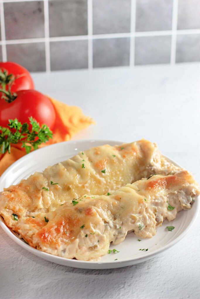 Two baked White Chicken Enchiladas topped with melted cheese and garnished with parsley, served on a white plate. Fresh tomatoes and parsley appear in the background.
