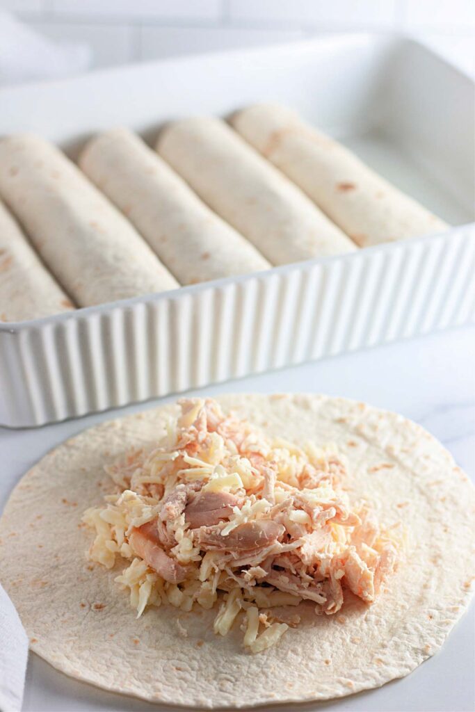 A flour tortilla filled with shredded chicken and cheese is being prepared in the foreground, perfect for making classic White Chicken Enchiladas, with rolled tortillas arranged in a white baking dish in the background.