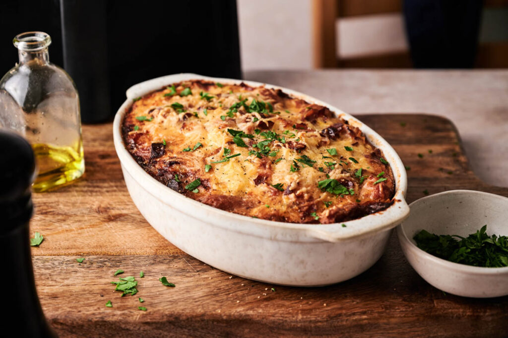 A baked lasagna in a white oval dish sits on a wooden board, garnished with chopped parsley, with a small bowl of extra parsley and a bottle of olive oil nearby—perfect alongside an Air Fryer Amish Breakfast Casserole for a hearty meal.