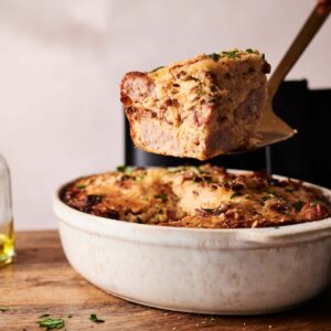 A hand lifts a slice of Air Fryer Amish Breakfast Casserole from a white baking dish on a wooden board, garnished with chopped herbs.