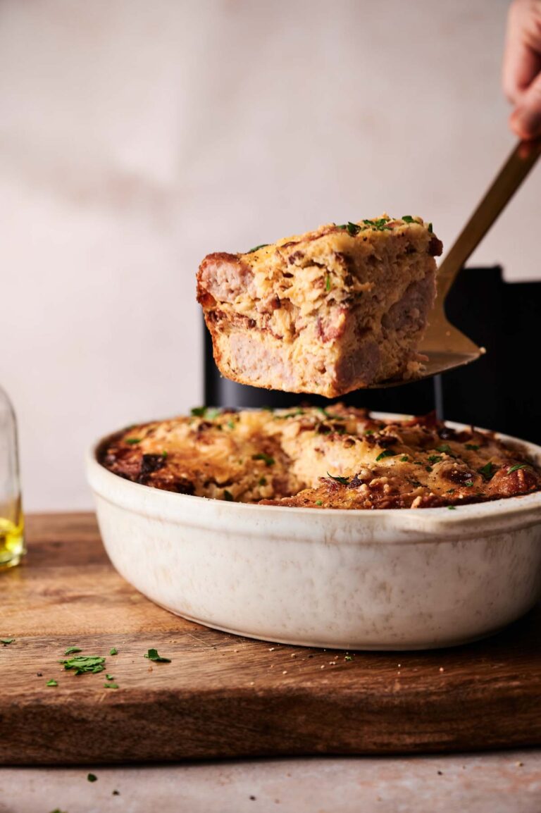 A hand lifts a slice of Air Fryer Amish Breakfast Casserole from a white baking dish on a wooden board, garnished with chopped herbs.