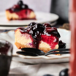 A slice of Air Fryer Blueberry Cheesecake topped with blueberry sauce sits on a plate, a spoon beside it and another slice in the background.