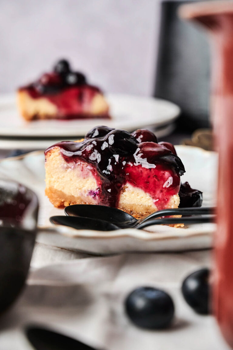 A slice of Air Fryer Blueberry Cheesecake topped with blueberry sauce sits on a plate, a spoon beside it and another slice in the background.