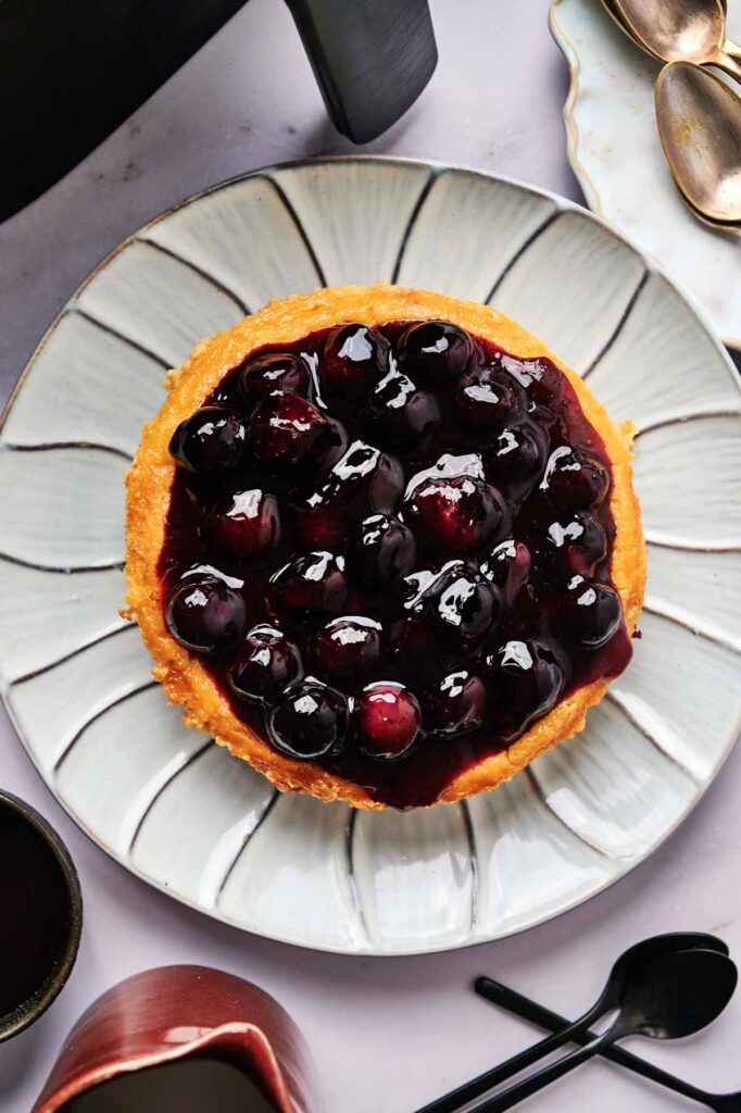 A round Air Fryer Blueberry Cheesecake topped with glossy blueberry sauce sits on a white plate, surrounded by utensils and dishes on a light-colored surface.